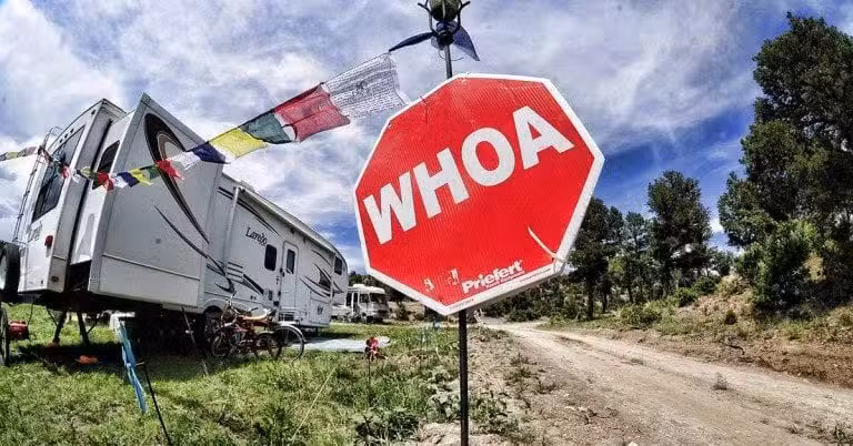 A red octagonal sign with the word WHOA stands beside a dirt road near a parked RV, bikes, and colorful flags, surrounded by trees and a partly cloudy sky.