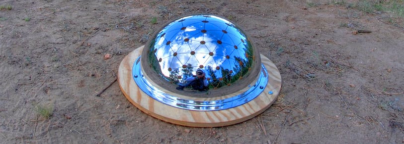 A shiny hemispherical dome made of reflective metal is mounted on a round wooden base and placed on bare, dry ground at Colorado Burning Man. The dome mirrors the sky, trees, and a person taking the photo in its gleaming surface.