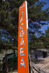 sign A tall, orange vertical sign with the word Apogaea in white capital letters stands outdoors. In the background, there are trees, a green vehicle, and a small structure under a blue sky.
