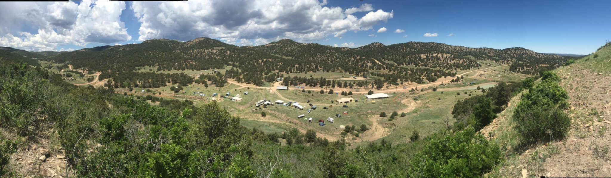 A panoramic view of a hilly landscape under a partly cloudy sky. Vegetation covers the foreground, with scattered trees and a cluster of tents and vehicles in an open area. Rolling hills stretch into the background.