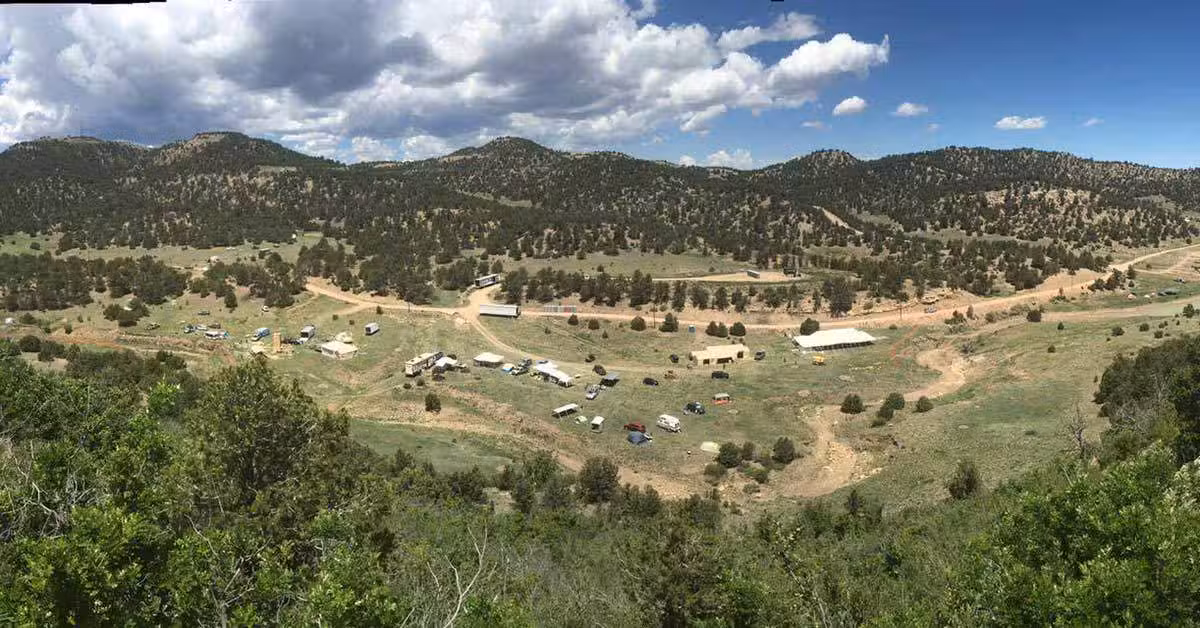 A panoramic view of a rural landscape with scattered trees, dirt roads, and small buildings. Tents and vehicles are visible across grassy fields. Rolling hills and a cloudy blue sky serve as the backdrop.