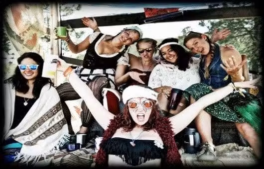 A group of six people are happily posing together. They are wearing casual, bohemian-style clothing and sunglasses, with one person in front enthusiastically spreading their arms wide. The setting appears to be outdoors.