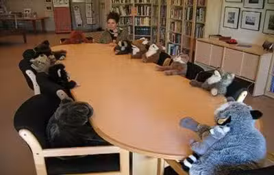 A person sits at a long conference table with numerous plush animal toys seated around it, resembling a formal meeting. Shelves filled with books line the walls in the background.