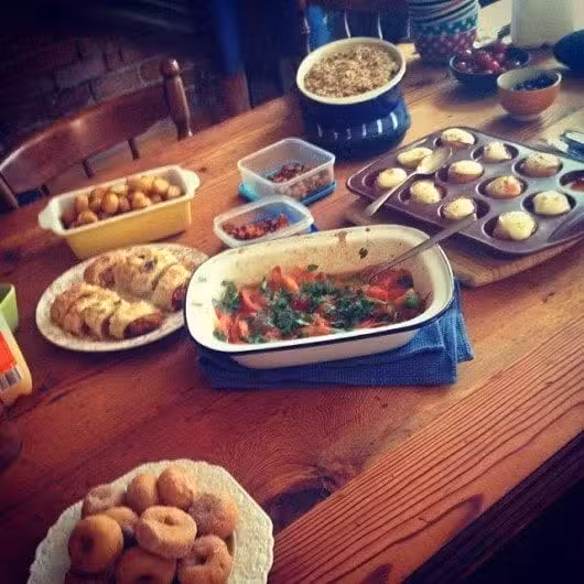 A wooden table is filled with a variety of dishes: a casserole, savory pastries, a platter of stuffed tomatoes, roasted potatoes, a dish of donuts, and multiple bowls and containers with various sides.