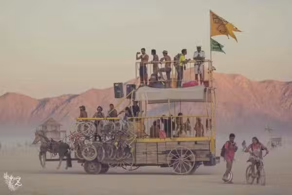 A multilevel vehicle adorned with bicycles transports people across a dusty desert reminiscent of the Colorado Burning Man event. Several individuals stand on top, next to flags. In the background, others ride bikes, with mountains visible under a hazy sky.