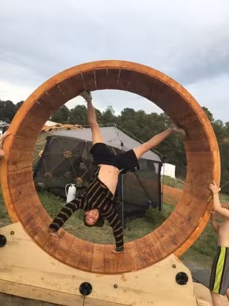 Amidst the vibrant landscape reminiscent of a Colorado Burning Man event, a person is doing a handstand inside a large wooden structure resembling a human-sized hamster wheel. With tents and trees in the background, two others assist by holding the wheel steady.