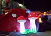 Inflatable red mushrooms with white spots and glowing stems create a whimsical scene outside a large red tent at night, reminiscent of a Colorado Burning Man event. Colorful wall murals adorn the background as people gather near the vibrant tent.