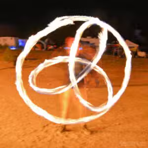 At a Colorado Burning Man event, a person performs a mesmerizing fire dance at night, spinning flames in overlapping circular patterns. The motion creates a glowing, dynamic display against the dark background, with blurry details suggesting movement and energy.