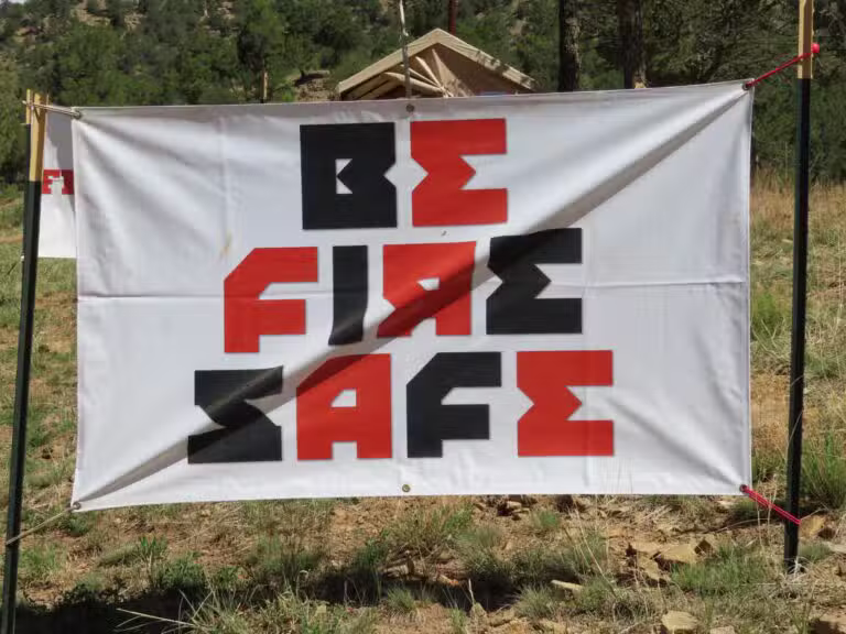 A large outdoor sign reads BE FIRE SAFE in bold, block letters with a mix of red and black colors. Set against a backdrop of trees and a tent, this scene echoes the spirit of a Colorado burner gathering in the wilderness.