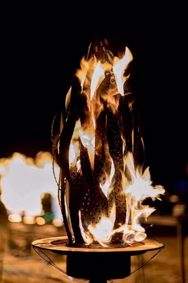 A sculptural fire pit, reminiscent of the creativity seen at the Colorado Burning Man event, crafted from a repurposed metal object—perhaps a washing machine drum. Flaming tendrils stretch skyward, casting a warm glow against the darkened backdrop.