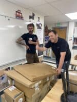 Two men smiling in a classroom stand behind a stack of cardboard boxes on tables. One man gives a thumbs-up while the other poses with a hand on his hip. Posters and classroom decorations are visible in the background.