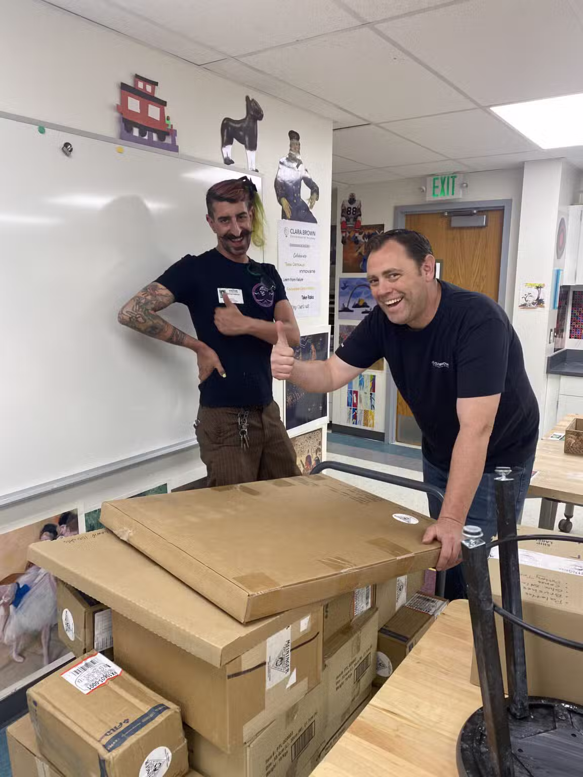 Two men stand smiling in a classroom, one giving a thumbs up, surrounded by large cardboard boxes on a table. The room has posters, art, and a whiteboard in the background.