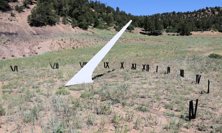 A large, white sundial gnomon stands in a grassy, open field surrounded by black Roman numeral hour markers, with trees and hills in the background under a clear blue sky.