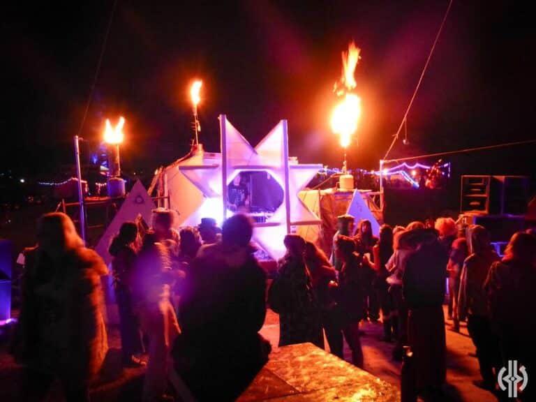 A crowd gathers at night in front of a brightly lit geometric stage with fire torches and speakers, creating a lively festival atmosphere under the dark sky.