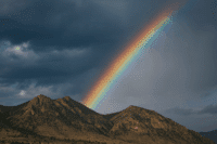 A vivid rainbow arcs across a dark, cloudy sky above rugged, rocky hills and patches of green vegetation.