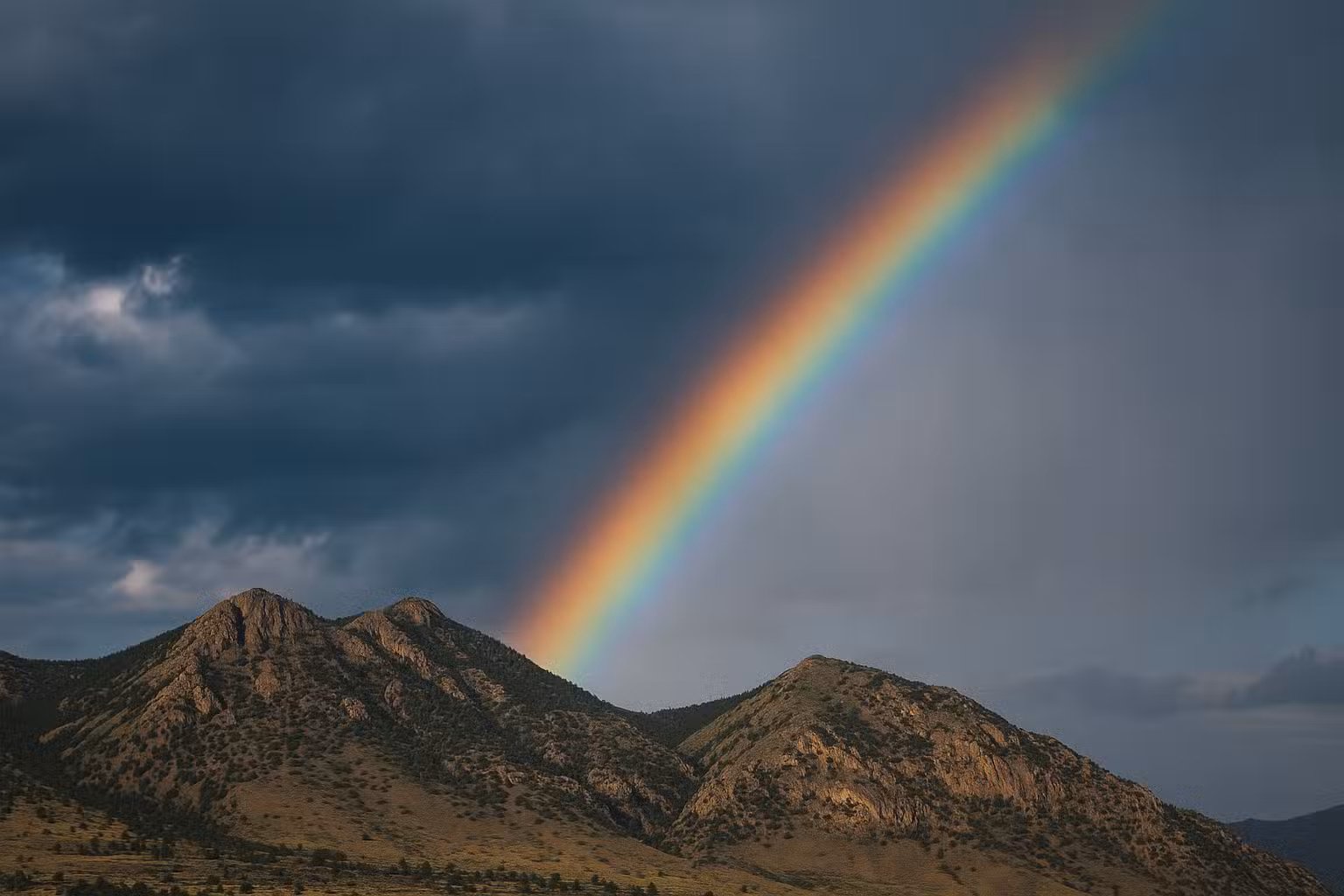 A vivid rainbow arcs across a dark, cloudy sky above rugged, rocky hills and patches of green vegetation.