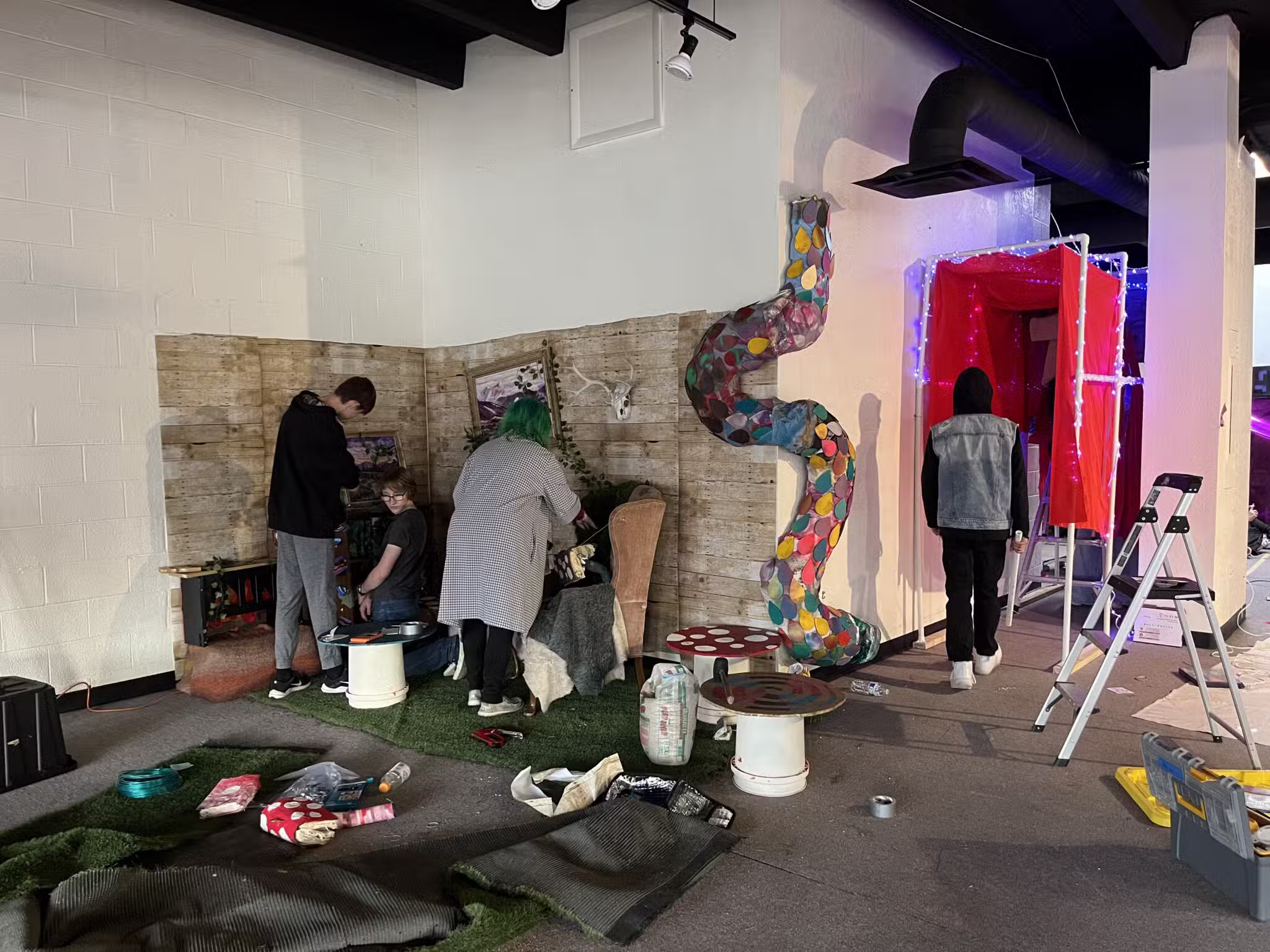 Several people work on setting up a whimsical indoor scene with artificial grass, a colorful twisting slide, and mushroom decorations. Art supplies and tools are scattered around, and two people stand near a lit-up doorway.