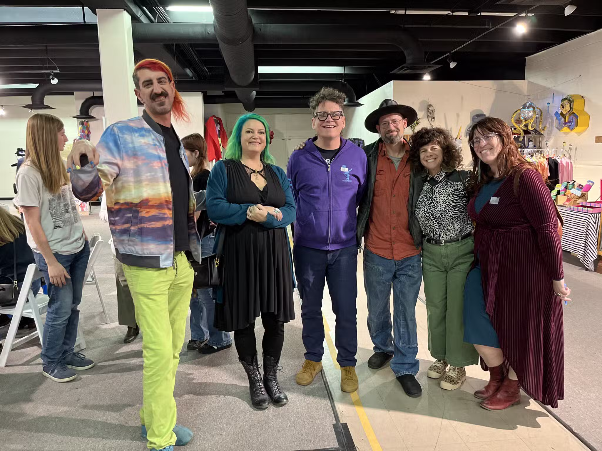 Six adults stand smiling together in an indoor art space, surrounded by colorful artwork and displays. The group wears casual, eclectic clothing, and the atmosphere appears lively and friendly.