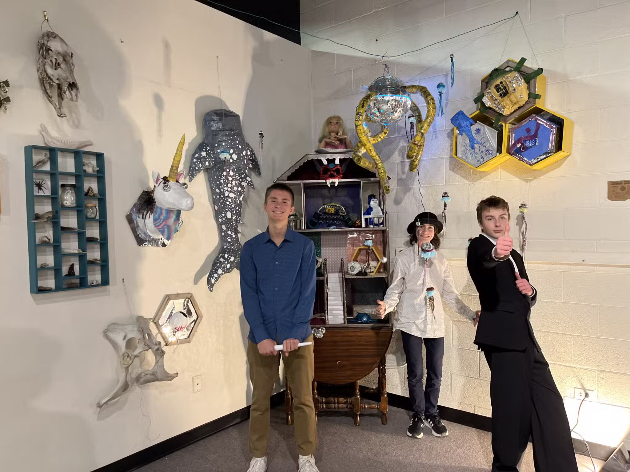 Three young people pose in front of a display of colorful, imaginative sculptures and art pieces on a white wall. Two smile cheerfully while one in a suit points dramatically toward the camera.