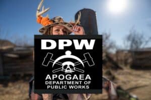 A person wearing a hat decorated with antlers and orange details stands outdoors, partly obscured by a large “DPW Apogaea Department of Public Works” sign with hammers and a skull logo. The background is blurred.