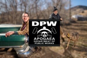 Two men are outdoors near an old car; one sits on the trunk smiling, the other walks a brown dog. A large DPW Apogaea Department of Public Works sign with a skull and wrenches covers the image’s center. Trees and hills are in the background.