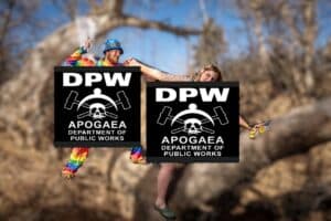 Two people in colorful outfits hold large signs reading DPW APOGAEA Department of Public Works with a skull, wrenches, and hard hat logo, standing outdoors with bare trees in the background.