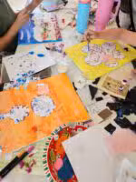 People painting colorful floral designs on paper surrounded by art supplies, sketches, glue, plastic cups, markers, and two pink water bottles on a table. Hands are actively drawing and painting in a creative setting.