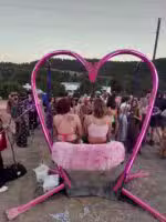 Two people sit on a pink, heart-shaped swing at an outdoor event. A crowd stands in the background, with trees and hills visible behind them. The scene has a festive, relaxed atmosphere.