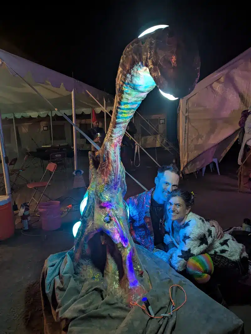 Arcturus, a tall iridescent creature sculpture with a glowing crescent head, lit at night beside two smiling participants.