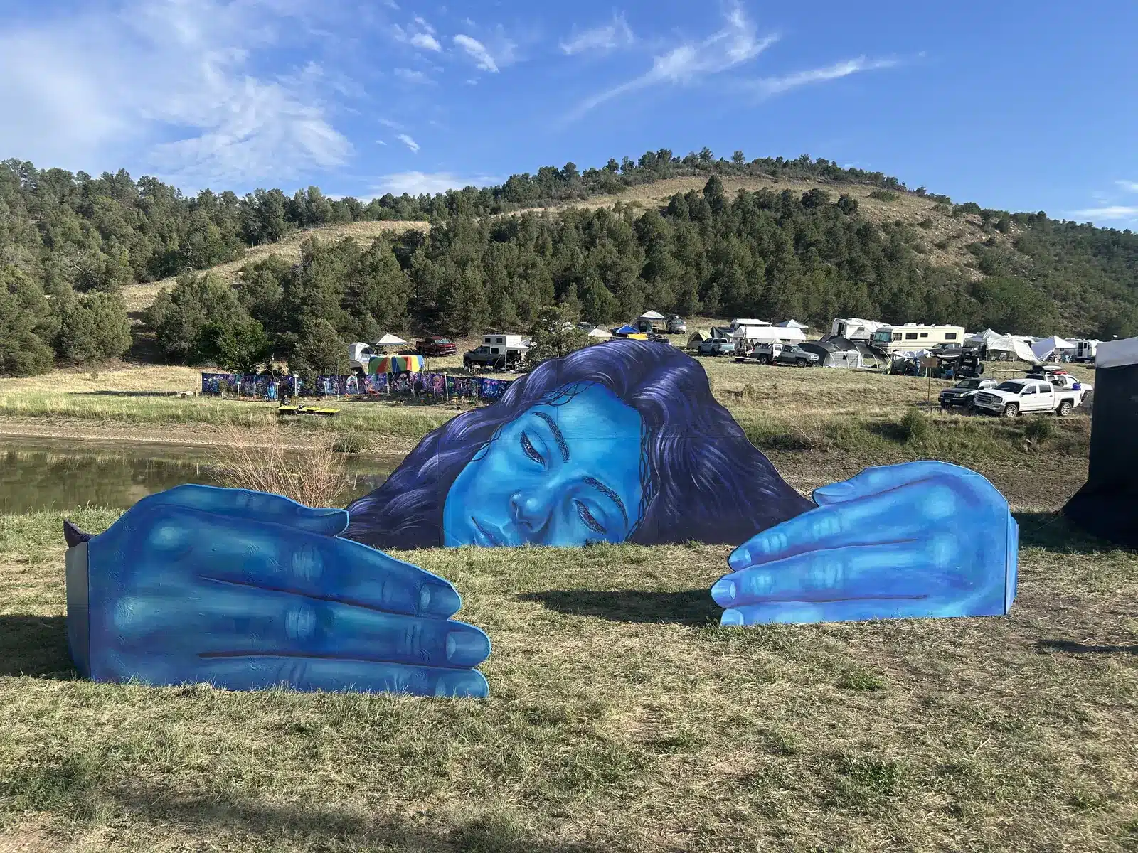 A giant blue painted face and two hands rising out of the grass at Apogaea 2026, with the campground and forested hills behind.