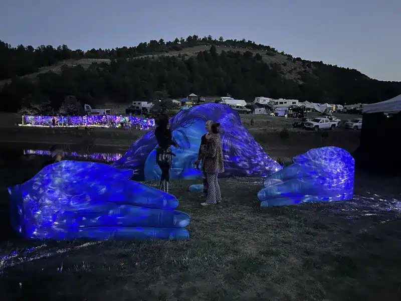 The giant blue face and hands sculpture glowing under blue light at dusk as visitors stand beside it.