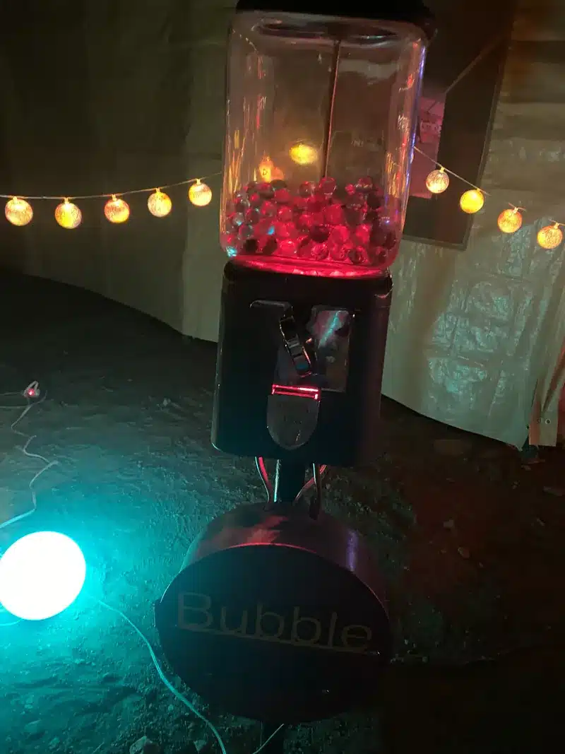 A lit gumball machine full of red orbs on a stand labeled Bubble, beneath a string of globe lights.