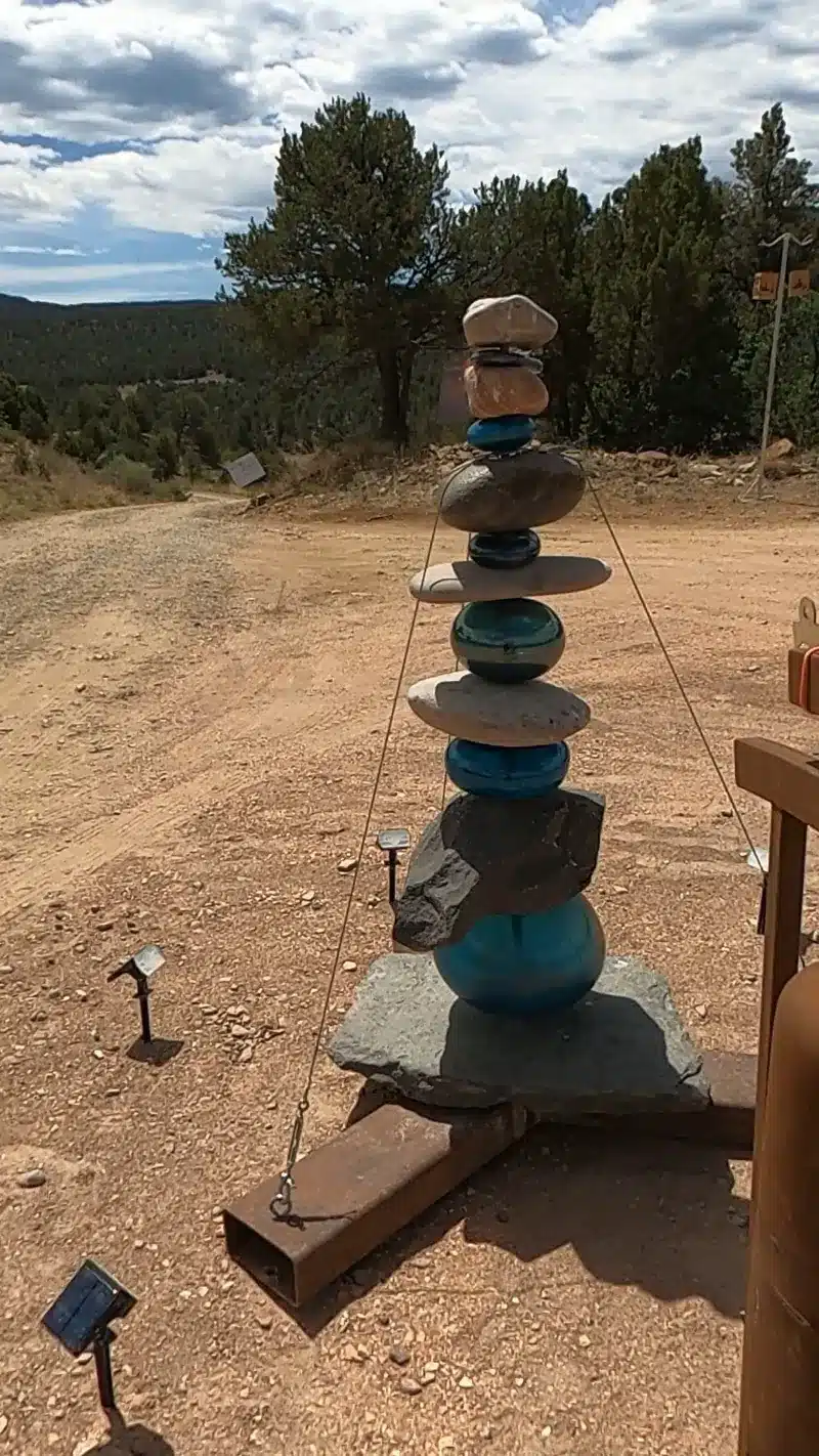 A tall cairn of stacked stones and blue glass orbs anchored on a steel base, with pines behind.