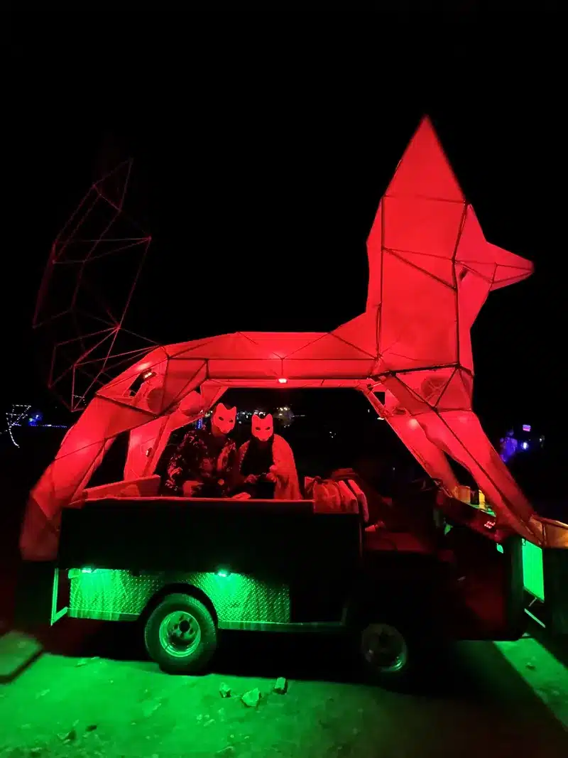 A geometric red fox art car glowing at night, with two masked riders seated inside.
