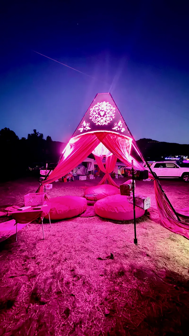 Love Letters, an A-frame shade structure with a laser-cut heart mandala, draped in pink fabric and glowing pink at night over floor cushions.