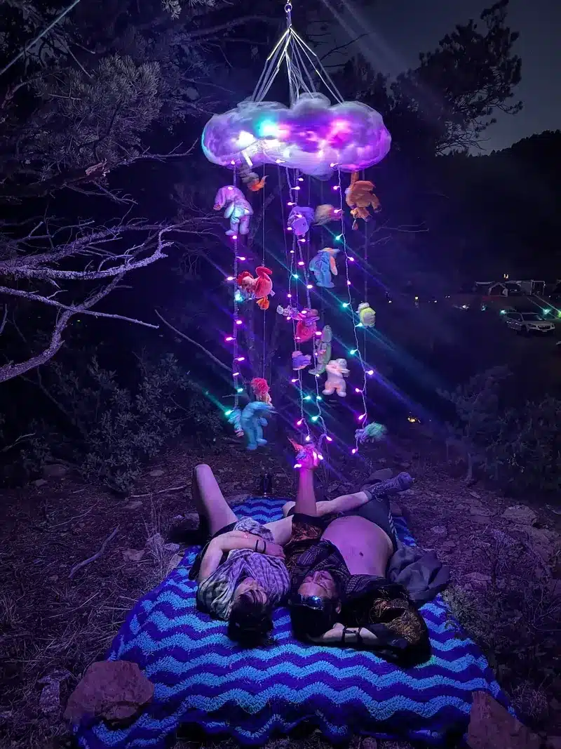 Nostalgia Mobile, a glowing cloud strung with lights and plush toys, hanging over two people lying on a blanket beneath it.