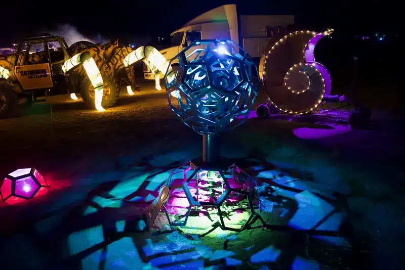 A cut-steel polyhedral sphere on a pedestal casting colorful shadow patterns at night, with a glowing nautilus-shell sculpture and animal lanterns behind it.