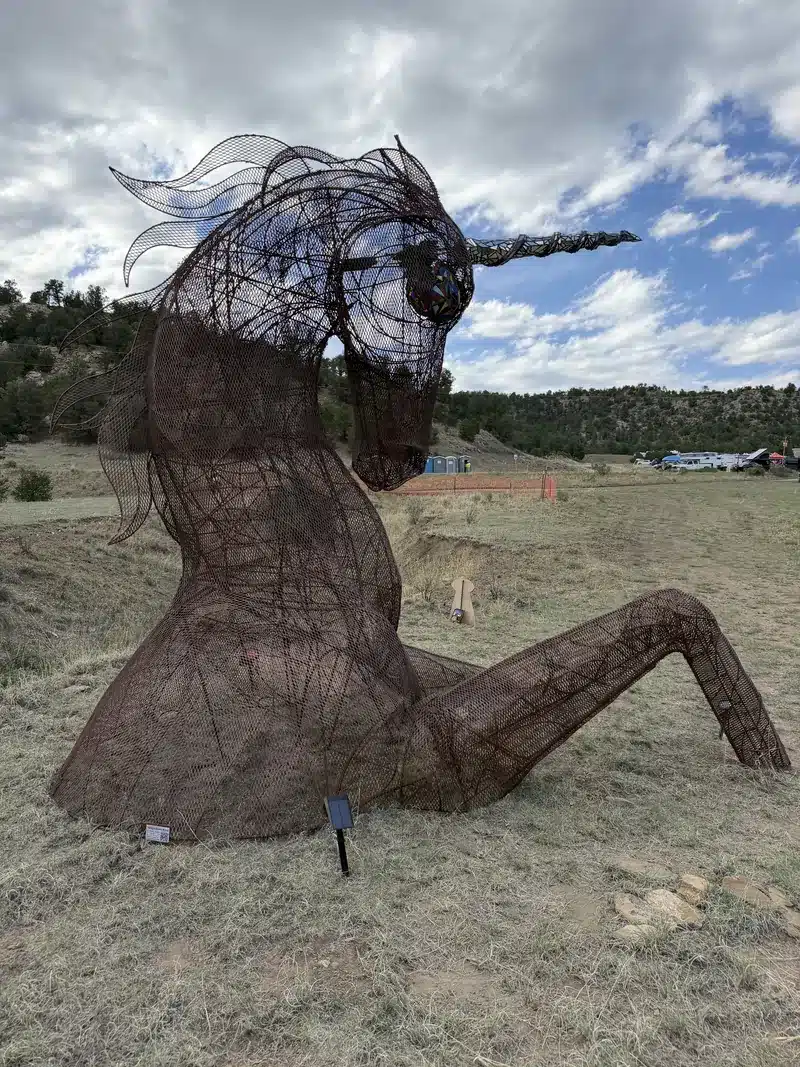 A giant rusted wire-mesh unicorn sculpture reclining in dry grass under a blue sky.