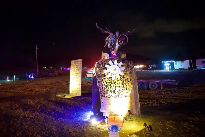 W.H.O.O., a cut-metal owl sculpture with a gear-pattern chest, glowing at night beside a laser-cut poem panel.