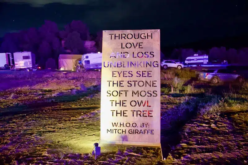 Laser-cut metal panel lit at night reading: Through love and loss unblinking eyes see the stone, soft moss, the owl, the tree. W.H.O.O. by: Mitch Giraffe.