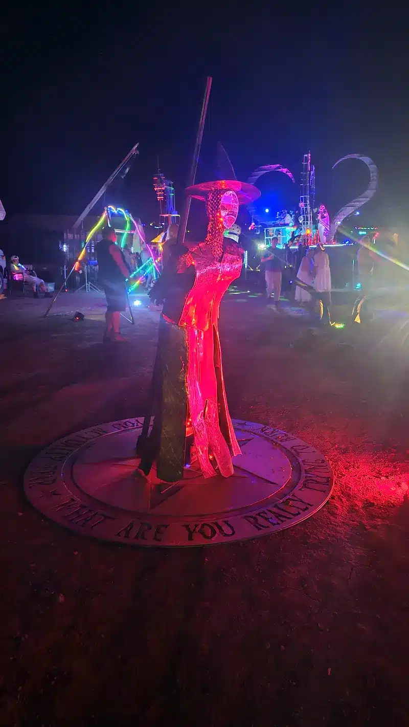 A metal witch sculpture glowing red at night, standing on a round plate engraved with the words Who are you really.