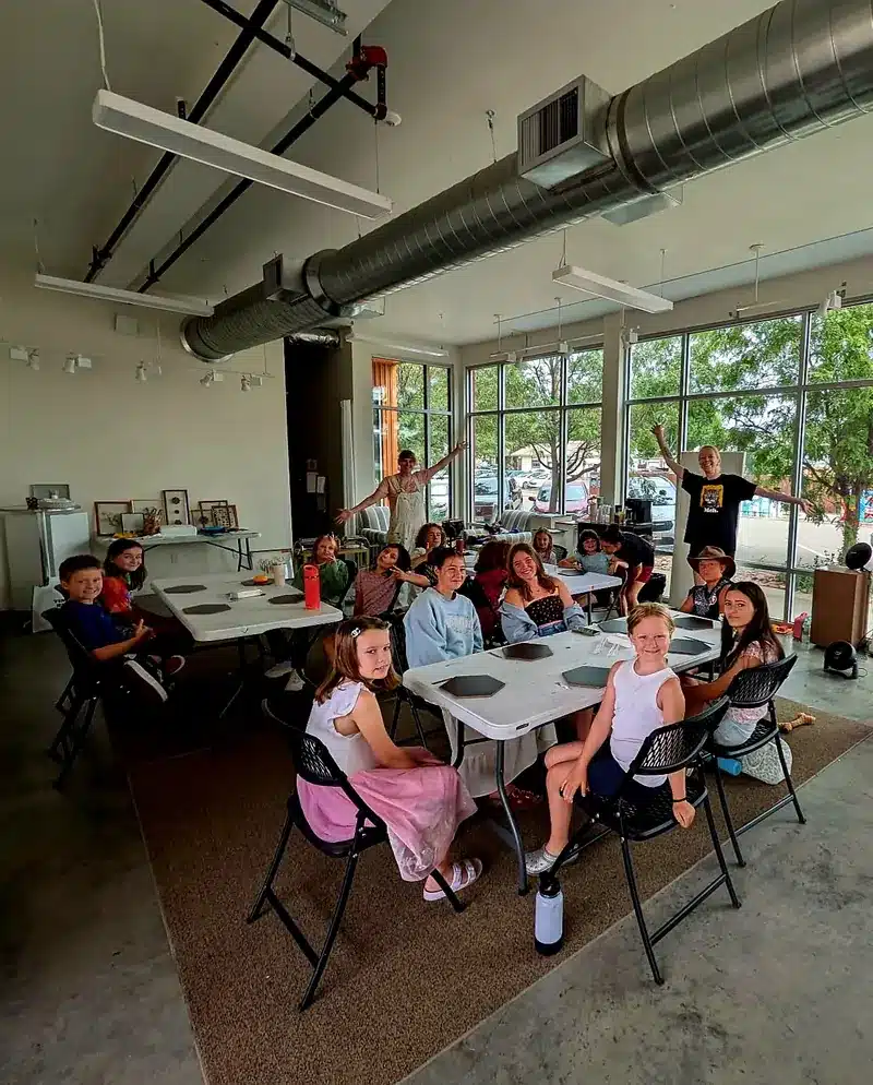 Kids at Monica Hsu's Loveland art camp seated around tables in a bright, airy studio, smiling at the camera as two instructors wave.