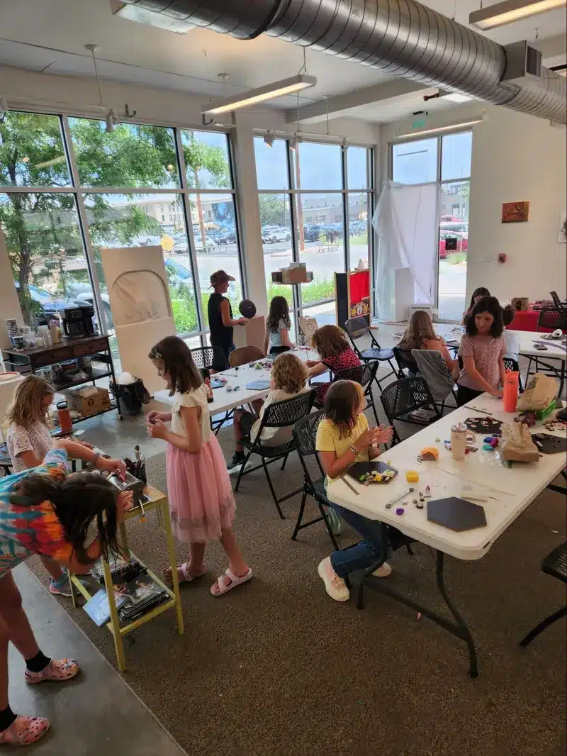 Campers working on art projects at tables and stations around the studio, with large windows looking out on trees.