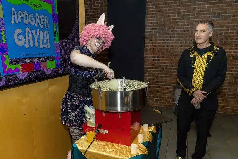 A guest in a pink curly wig and bunny ears spins cotton candy at last year's Apogaea Gayla, beside a colorful Gayla sign, as another guest looks on.