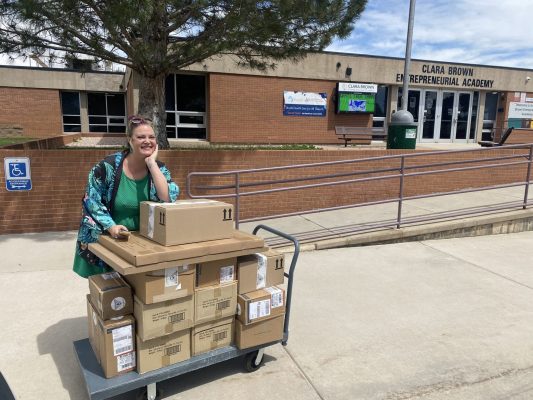 A smiling woman stands outside a building labeled Clara Brown Entrepreneurial Academy, leaning on a cart stacked with several cardboard boxes on a sunny day. A smiling woman stands outside a building labeled Clara Brown Entrepreneurial Academy, leaning on a cart stacked with several cardboard boxes on a sunny day.