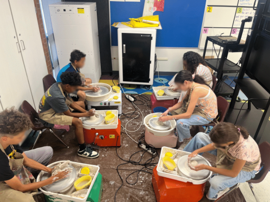 Six children sit around pottery wheels in a classroom, shaping clay with their hands. Yellow sponges and containers of water are on their tables. The floor is messy with clay residue. The students appear focused on their work.