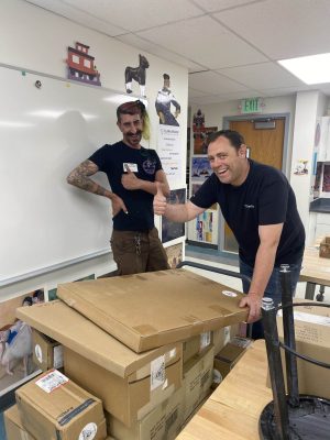 Two men smiling in a classroom stand behind a stack of cardboard boxes on tables. One man gives a thumbs-up while the other poses with a hand on his hip. Posters and classroom decorations are visible in the background. Two men smiling in a classroom stand behind a stack of cardboard boxes on tables. One man gives a thumbs-up while the other poses with a hand on his hip. Posters and classroom decorations are visible in the background.