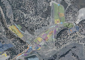 A satellite map of a rural area with trees, roads, and parking lots, overlaid with various colored sections indicating different zones or plots. Several icons mark points of interest along the roads.